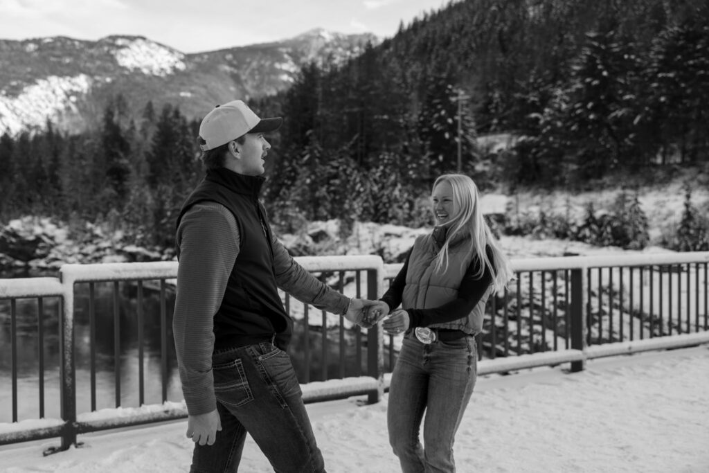 Black and white photo of an engaged couple walking hand in hand across Belton Bridge near the West Glacier entrance to Glacier National Park — Lindsey and Ethan — Glacier National Park Winter Engagement — Stacy Edwards Photography
