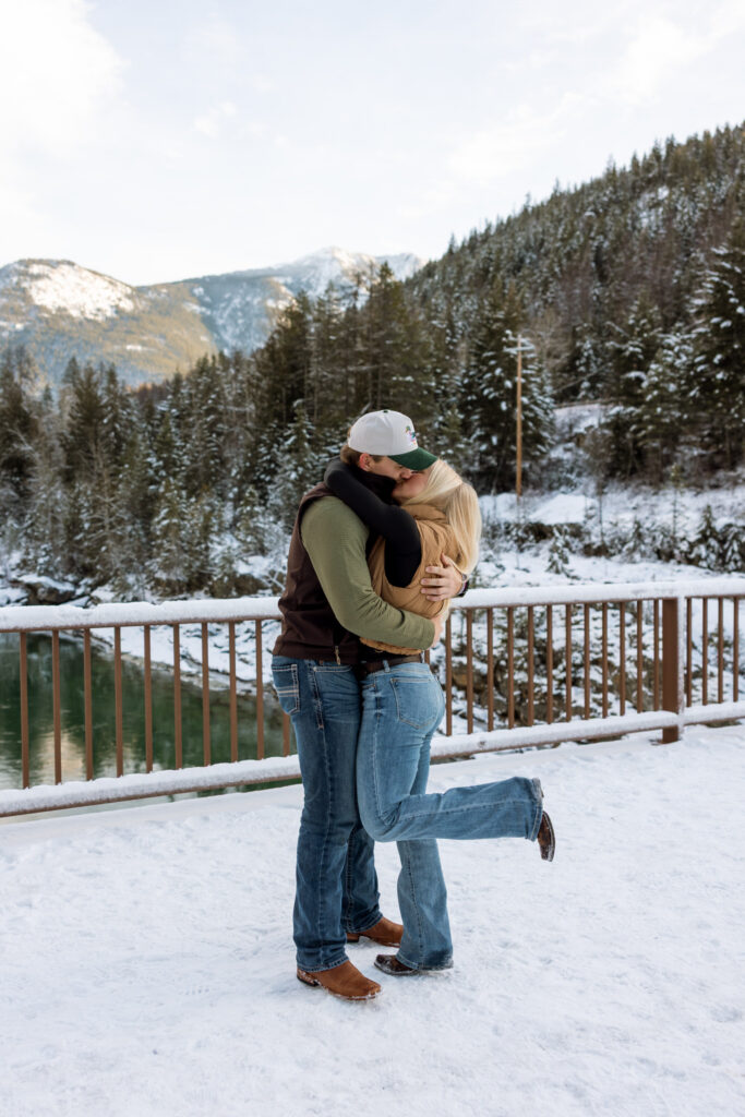 Engaged couple sharing a kiss on Belton Bridge surrounded by snow-covered evergreen trees near West Glacier — Lindsey and Ethan — Glacier National Park Engagement Photographer — Stacy Edwards Photography