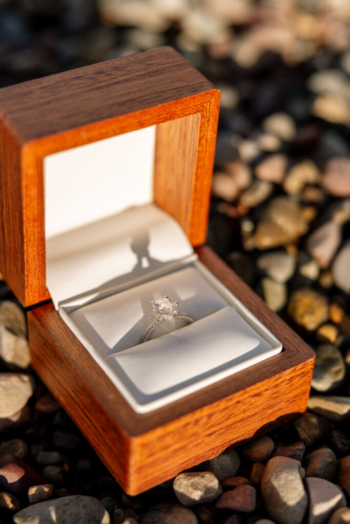 Diamond engagement ring in a wooden ring box resting on the rocky shore of Lake McDonald — Lindsey and Ethan — Sandy Point Glacier National Park Proposal — Stacy Edwards Photography