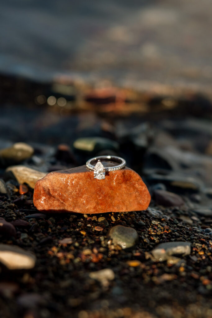 Pear cut diamond engagement ring resting on a red rock at the shore of Lake McDonald — Lindsey and Ethan — Sandy Point 7 Mile Pullout Glacier National Park — Stacy Edwards Photography