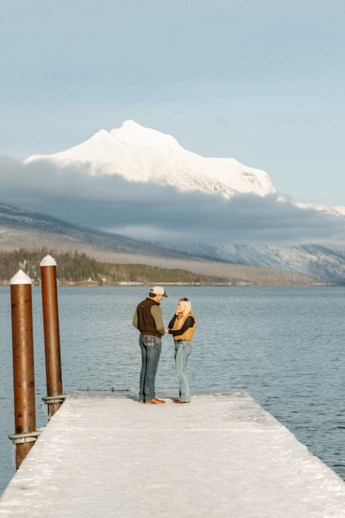 Couple sharing a surprised reaction moment on a snow-covered dock at Apgar Village — Lindsey and Ethan — Lake McDonald Glacier National Park Winter Proposal — Stacy Edwards Photography