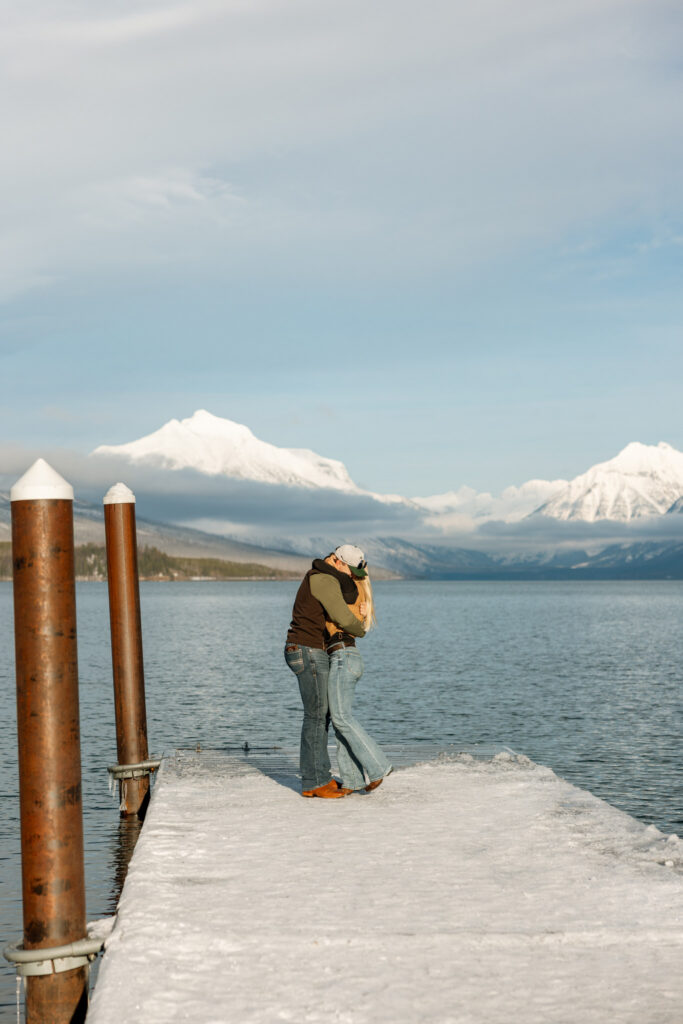 Newly engaged couple sharing their first kiss on a snowy dock at Lake McDonald — Lindsey and Ethan — Apgar Village Glacier National Park Proposal — Stacy Edwards Photography