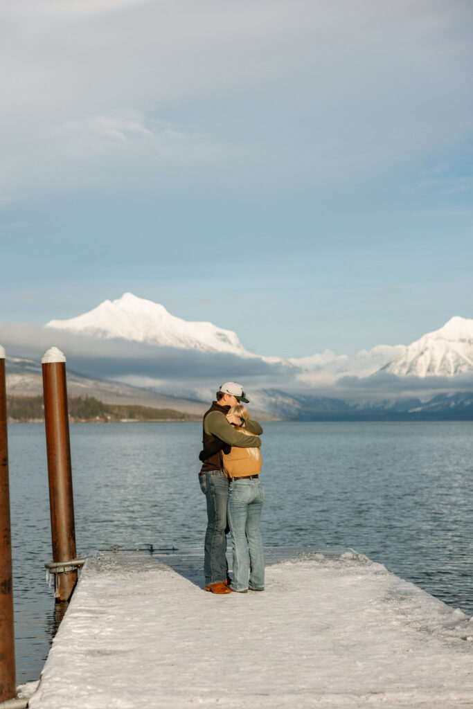 Newly engaged couple embracing on the Apgar Village dock with snow-capped Glacier National Park mountains reflected in Lake McDonald — Lindsey and Ethan — Stacy Edwards Photography