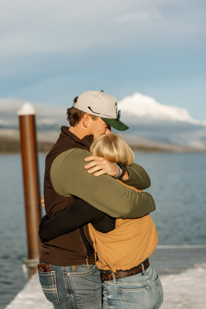 Man kissing his newly engaged partner on the forehead on a snowy dock at Lake McDonald — Lindsey and Ethan — Glacier National Park Winter Proposal — Stacy Edwards Photography