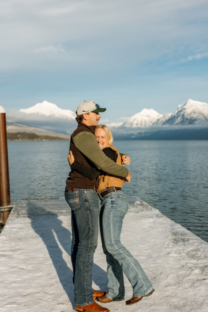 Newly engaged couple laughing and hugging on a snow-covered dock at Apgar Village — Lindsey and Ethan — Lake McDonald Glacier National Park — Stacy Edwards Photography