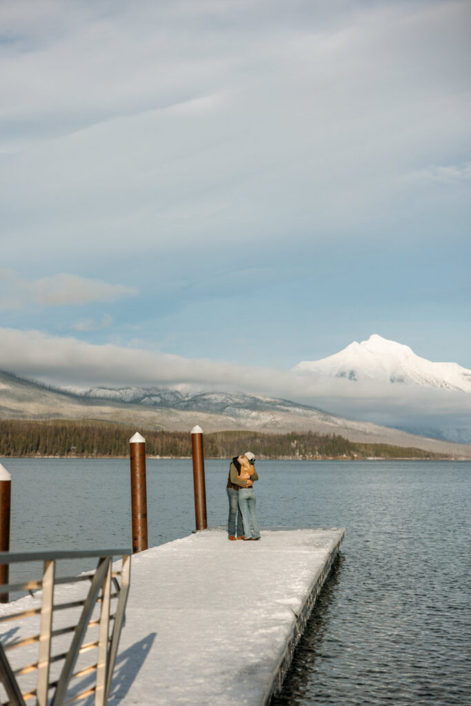 Newly engaged couple embracing at the end of the Apgar Village dock with Glacier National Park mountains in the background — Lindsey and Ethan — Lake McDonald Winter Proposal — Stacy Edwards Photography
