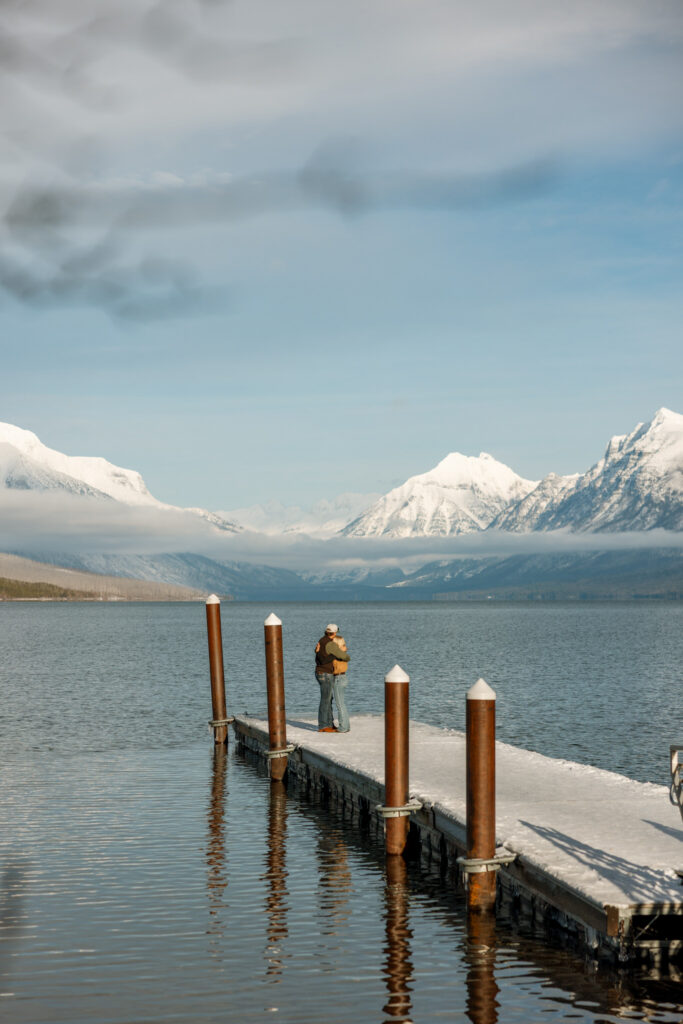 Engaged couple standing together on a snow-covered dock with sweeping Lake McDonald and Glacier National Park mountain views — Lindsey and Ethan — Apgar Village Glacier National Park Proposal Photographer — Stacy Edwards Photography