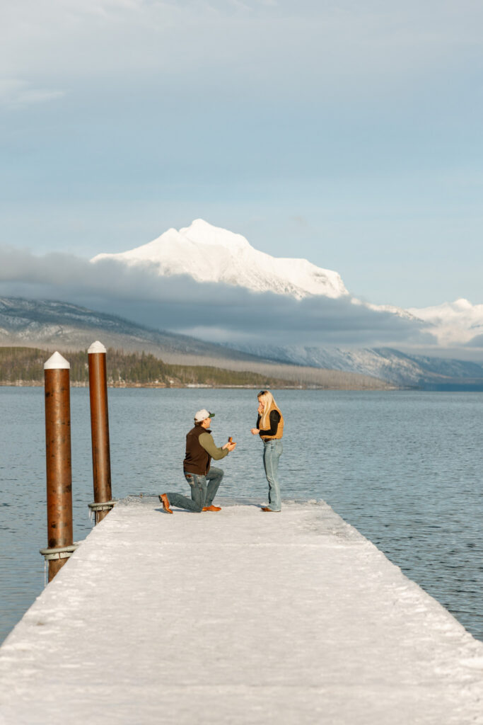 Man on one knee proposing on a snow-covered dock with snowy mountain backdrop — Lindsey and Ethan — Apgar Village Lake McDonald Glacier National Park — Stacy Edwards Photography