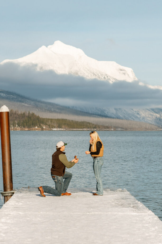 Woman covering her mouth in surprise as her partner proposes on a snowy Lake McDonald dock — Lindsey and Ethan — Glacier National Park Winter Proposal — Stacy Edwards Photography