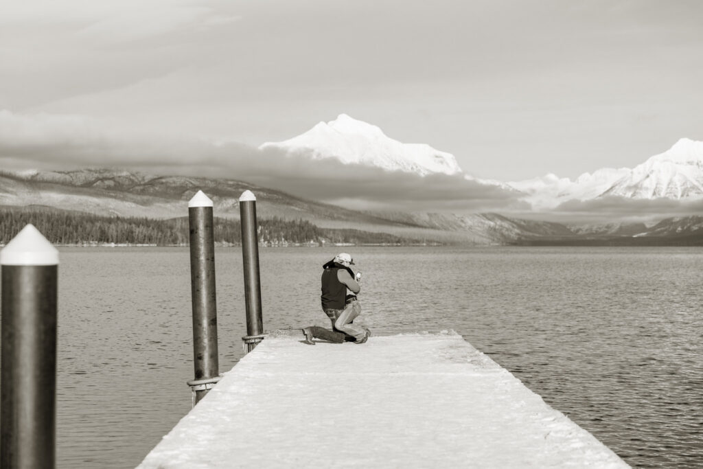 Black and white image of a man on one knee proposing on the Apgar dock at Lake McDonald — Lindsey and Ethan — Glacier National Park Winter Proposal — Stacy Edwards Photography
