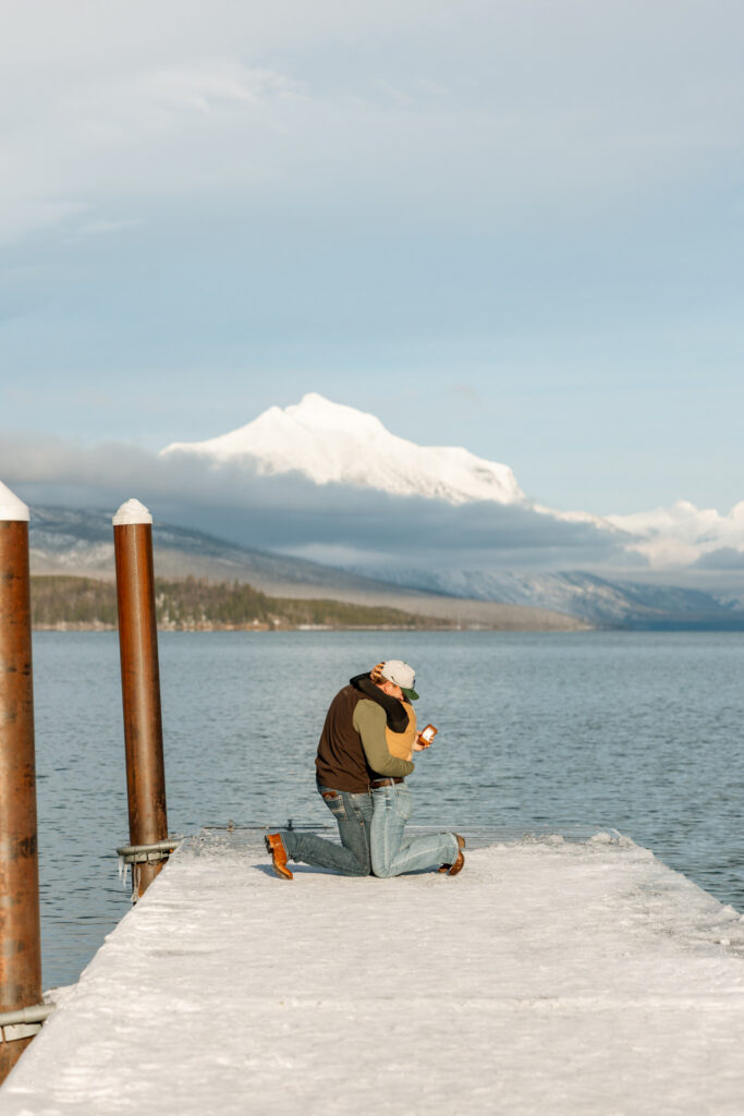 Man kneeling on a snowy dock holding an open ring box at Lake McDonald Glacier National Park — Lindsey and Ethan — Glacier National Park Proposal Photographer — Stacy Edwards Photography